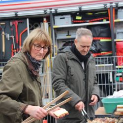 Ein Man steht am Grill und wendet die Bratwürste - eine Frau steht daneben mit einer Holzzange und einem Stück Toast.