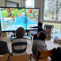 Eine Gruppe von Kindern sitzt vor einer Digitalen Tafel während die Lehrerin an der Tafel etwas erklärt.