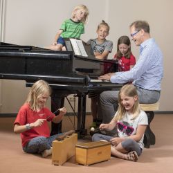 Der Lehrer Sitzt am Piano. Zwei Schülerinnen sitzen vor ihm und spielen das Xylophone. Hinter ihm stehen zwei Schülerinnen. Und auf dem Piano sitzt eine Schülerin.