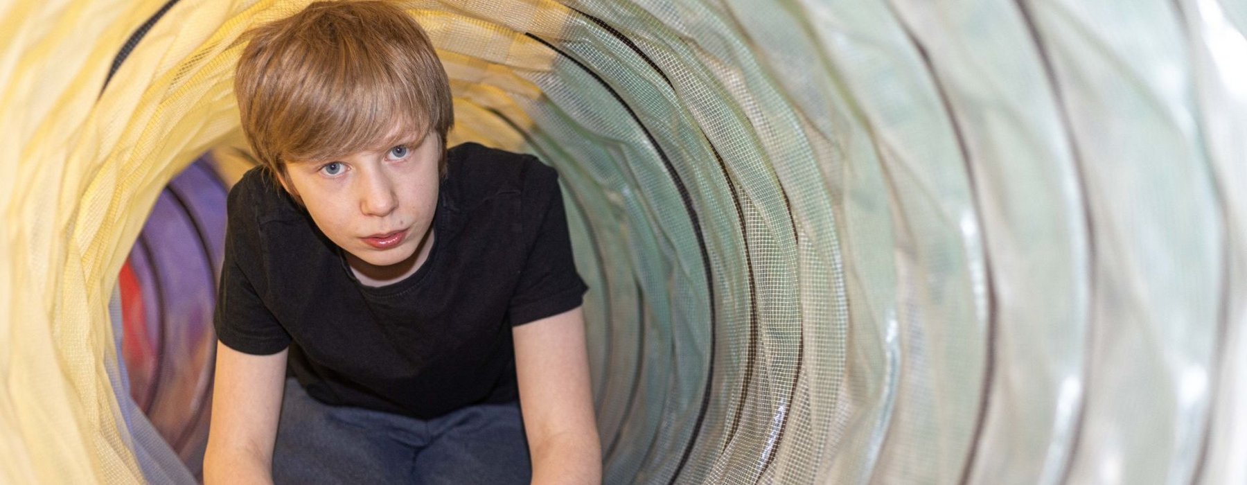 Ein Blonder Junge grabbelt durch eine Spielröhre und schaut direkt in die Kamera. Die Kamera schaut von einem ende der Spielröhre rein.