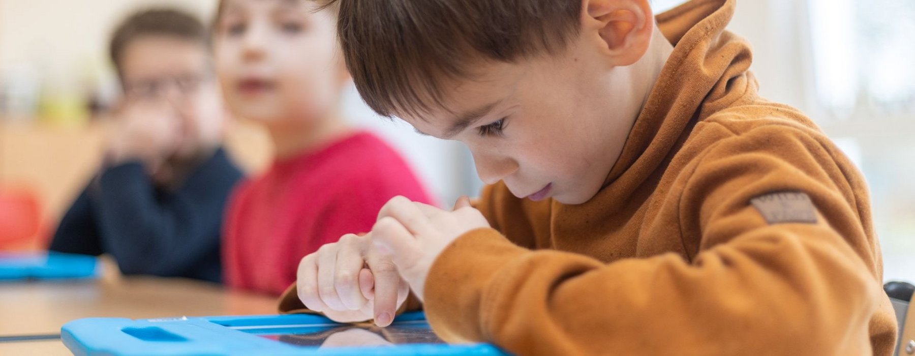 Ein Junge ist im Fokus der an einem Lerntablet sitzt nicht im Fokus sind zwei andere Kinder