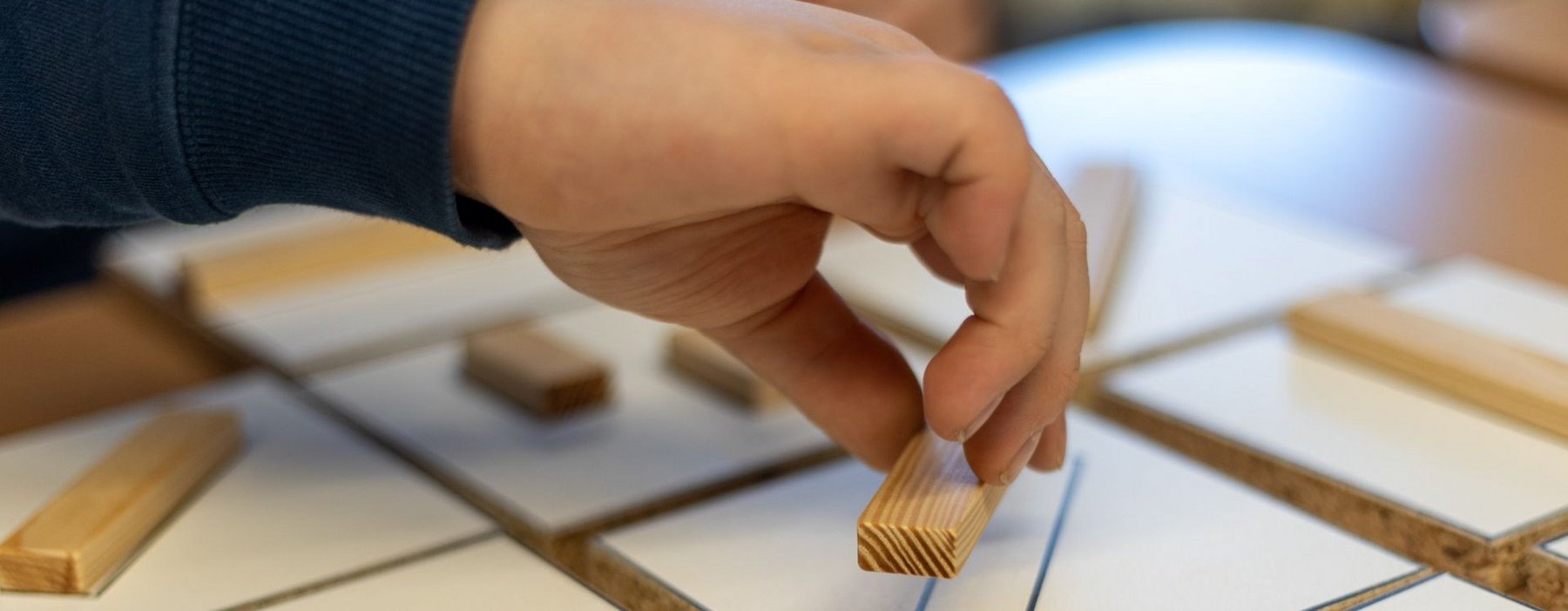 Eine Hand platziert einen Kleinen Handgroßen Holzbalken in eine aufgemalte Umrandung.